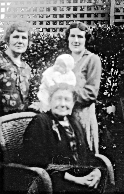 three women and a baby in a garden. Two standing, one seated. One of the standing women is holding a baby