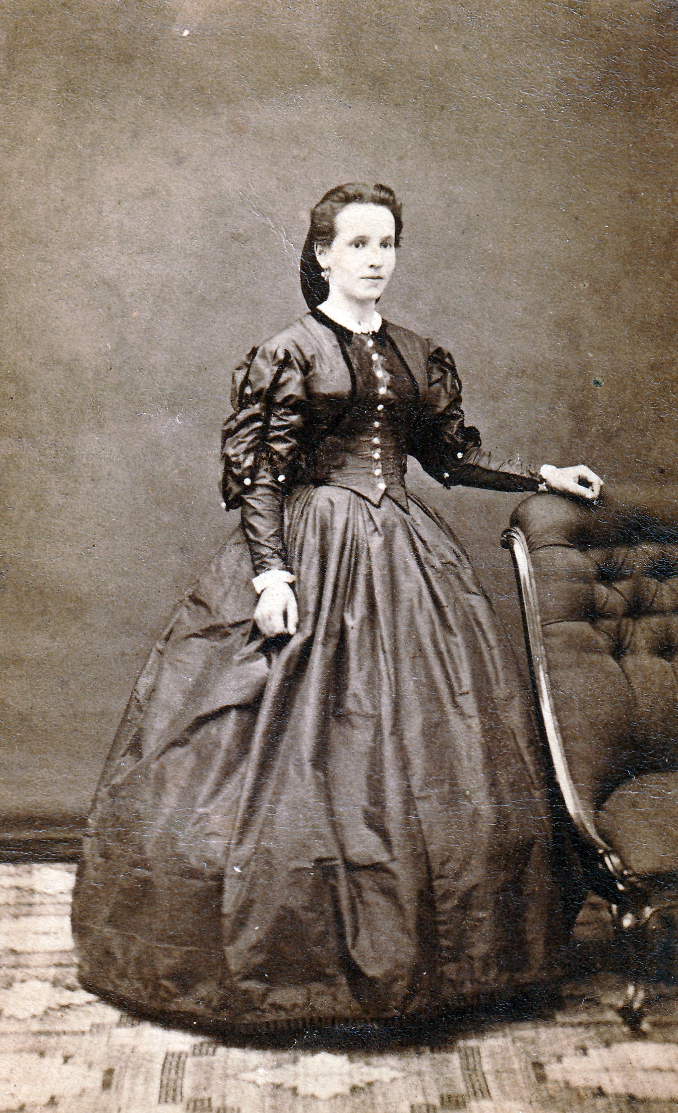 Studio portrait of a woman in dark dress with white buttons down the front. her right hand is resting on a dark chair