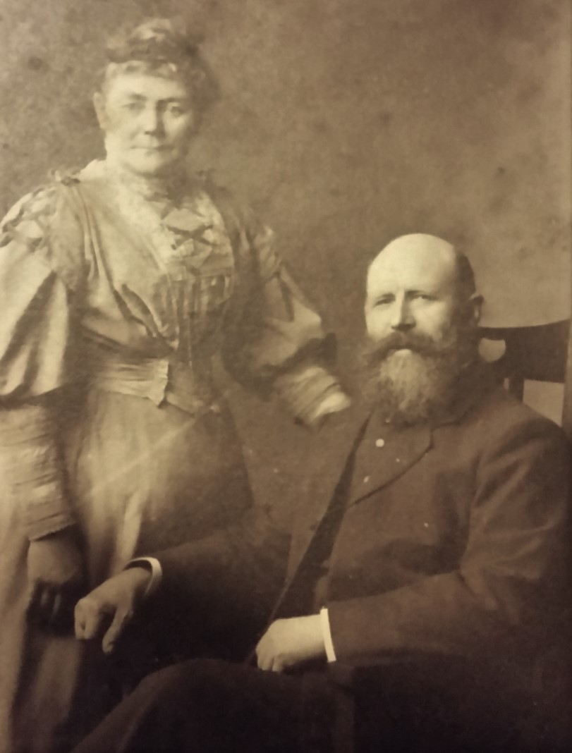 Portrait of a middle aged man and woman. The woman is standing and has hair in a bun on top of her head and a long sleeved dress on. The man is seated and is wearing a dark suit and has a dark beard. 