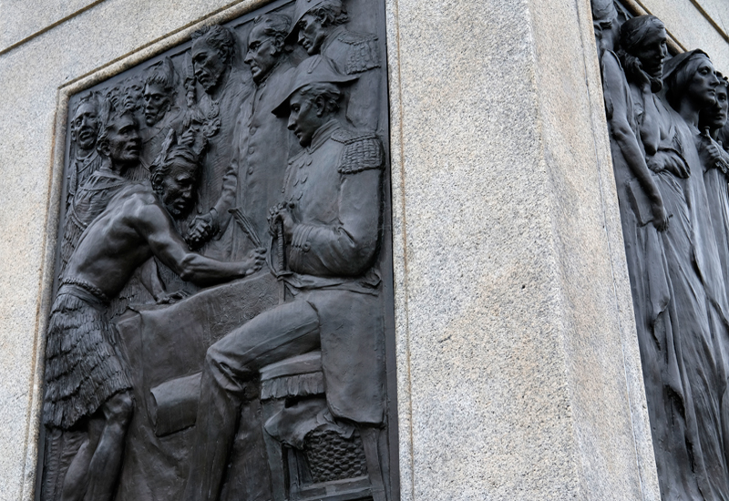 Details of bronze reliefs on Queen Victoria statue showing Māori and European figures signing document on one panel and a row of women on another.