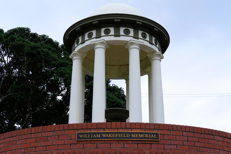 Round memorial with four pillars visible behind plaque with William Wakefield memorial inscription on red brick wall.