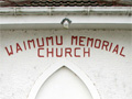 View of the entrance of the church. On the wall are two black plaques below an inscription in red letters that reads: 'Waimumu Memorial Church