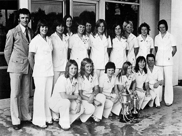 Sixteen members of a football team, all women, standing and crouching in two rows, plus one man. The women are all wearing white trouser suits. In front of them are two silvers cups decorated with ribbons.