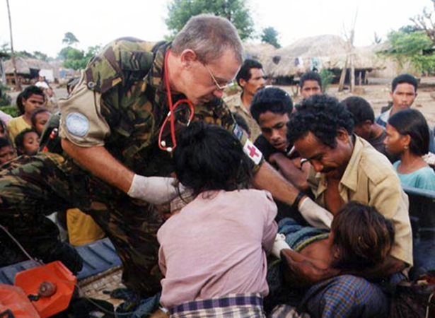 Man in camouflage military uniform wearing medical gloves and stethoscope surrounded by people in civilian clothing