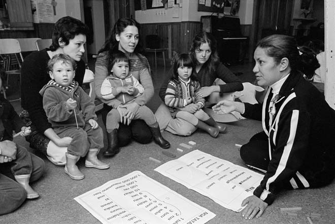 Group of women, some holding children, kneel on the floor looking at sheets of paper with te reo Māori words on them.