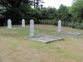 Group of white headstones on grass area surrounded by trees and bush.