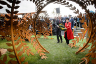 Two people approach steel sculpture holding a wreath of flowers.