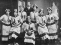 Seven women standing and seated in football uniforms, including long striped dresses and caps, one holding a football. Embroidered onto their dresses and printed on the ball is the word Polytechnic. Standing with them is a man in a suit. 