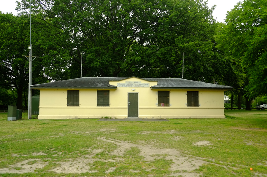 Exterior view of yellow and green building with patchy grass in the foreground and trees in the background
