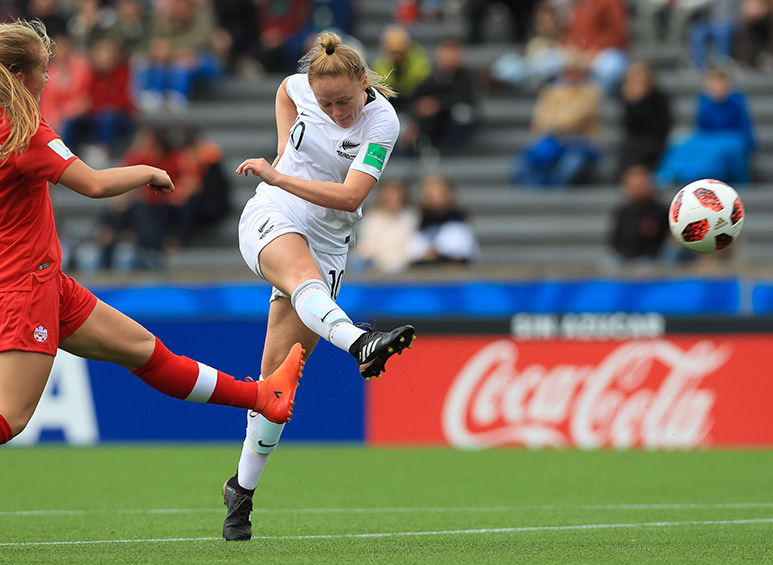 Woman in white uniform in the process of kicking a football, while a woman player in red uniform attempts to tackle her.