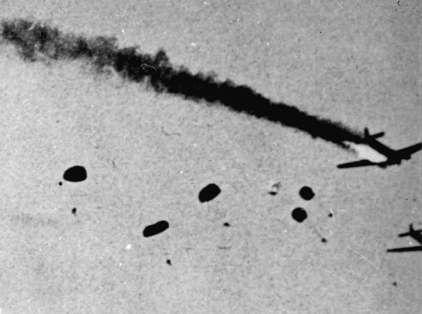 Plane with smoke pouring out the back surrounded by parachutes falling from the sky