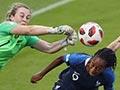 A young woman in green New Zealand goalkeeper uniform leaps into the air while reaching to push a ball away from a goal during a football game. A woman in a blue football uniform is partly crouched in front of her to avoid the punch, while other players in white New Zealand uniforms run towards her.