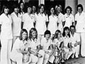 Group of women in matching uniforms, and one man in a suit, standing and crouching behind two trophies.