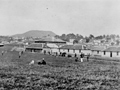 Historical image showing people playing cricket of a field in front of a group of buildings.