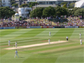 Men in whites playing cricket surrounded by crowd on grassy banks