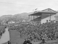 Historical photo of crowded banks and stand at cricket match with two vintage cars in the foreground