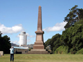 Two people looking at column monument on raised grassy area with silo building in background