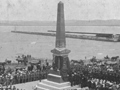 Memorial draped with NZ flags and large crowd gathered, including military personnel with heads bowed
