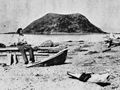 Man sitting on remains of a dinghy on beach with island in the background
