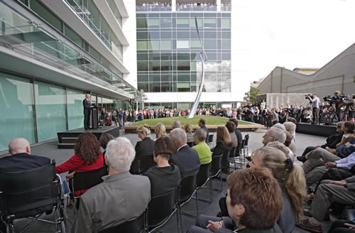 Seated audience listening to speaker next to memorial on lawn of modern building.