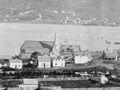 Historic image of Wellington showing Church standing out among scattered buildings, set with harbour in the background