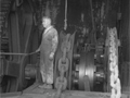 Man in dungarees next to large grinding wheel turing  pulleys with large chains