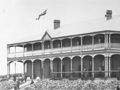 Historic photo of grand tw0-storey wooden building with a Union Jack flying above the entrance and set against a newly planted garden,