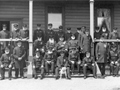 Group of elderly men, most with white beards and in uniforms with medals, posing for photo on steps of wooden building. A dog is held by one of the men sitting in the middle.