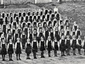 Side-by-side images, one showing rows of girls standing to attention in front of flag pole and the other showing students gathered around flagpole in front of school
