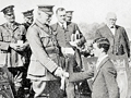 A crowd around a large shield on stage with high ranked soldier shaking hand of cadet
