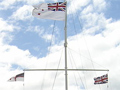 Flagpole wiht Navy Ensign, Union Jack and United Tribes flags set on large lawn overlooking the sea. Men in navy uniforms are gather around the base.