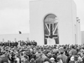 Opening of building with large crowd gathered in front of stage and building with Union Jack draped over entrance.