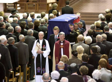 Coffin covered with New Zealand flag carried by men in uniform watched by people standing in church pews