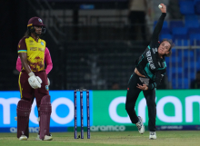 Person in black and teal sports uniform bowling a white ball next a set of cricket stumps and a person in a maroon and yellow sports uniform holding a wooden bat