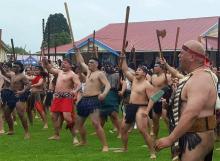 Group of bare-chested Māori men and boys carrying taiaha staffs with arms raised