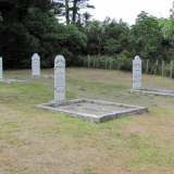 Group of white headstones on grass area surrounded by trees and bush