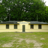 Exterior view of yellow and green building with patchy grass in the foreground and trees in the background