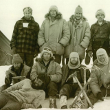 Group of ten men dressed up warmly posing for a photograph on the ice. One man lies in front while the others stand or kneel. Four ice picks are stuck in the ice in front of the group.