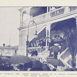 Historic image with crowd gathered on deck and grounds of two-storey wooden building with a lamp in front. One person is speaking to the crowdd