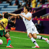 A woman in a New Zealand football uniform and player in a Cameroon uniform vie for the ball during a game.  