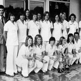 Sixteen members of a football team, all women, standing and crouching in two rows, plus one man. The women are all wearing white trouser suits. In front of them are two silvers cups decorated with ribbons.