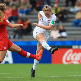 Woman in white uniform in the process of kicking a football, while a woman player in red uniform attempts to tackle her.