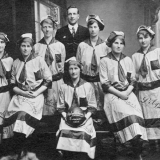 Seven women standing and seated in football uniforms, including long striped dresses and caps, one holding a football. Embroidered onto their dresses and printed on the ball is the word Polytechnic. Standing with them is a man in a suit. 
