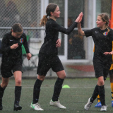 Three girls dressed in black football uniforms, playing football in the rain, high-five each other.