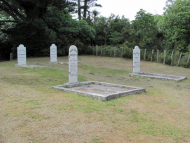 Group of white headstones on grass area surrounded by trees and bush