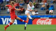 Woman in white uniform in the process of kicking a football, while a woman player in red uniform attempts to tackle her.