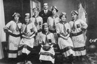 Seven women standing and seated in football uniforms, including long striped dresses and caps, one holding a football. Embroidered onto their dresses and printed on the ball is the word Polytechnic. Standing with them is a man in a suit. 