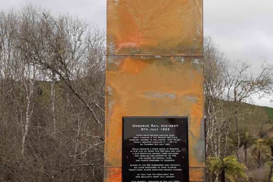 Bronze coloured pillar inscribed with date in te reo Māori with flowers laid around the base 