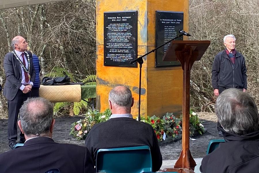 Two men standing solemnly either side of bronze pillar with people sitting in chairs and lectern and microphone in the foreground 