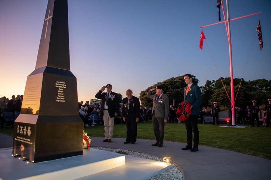 Four men wearing military medals and carrying floral wreath stand in front of black granite obelisk watched by crowd of people
